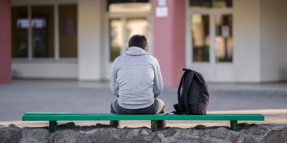 One young man sitting on bench at school yard. Break time. Back view.